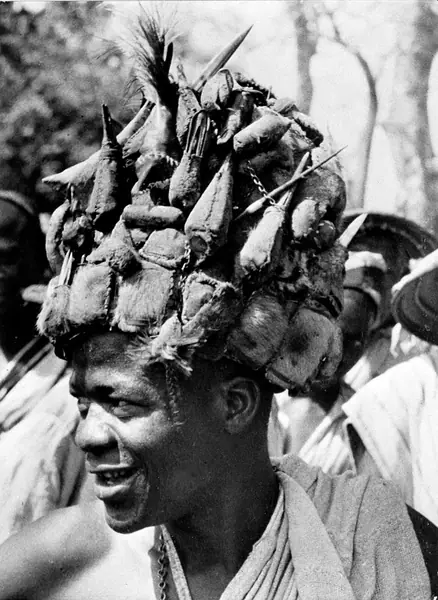 A West African witchdoctor with a hat entirely covered with amulets of leather and feathers. The leather is sewn into little pouches, each of which contains verses from the Koran and other charms.