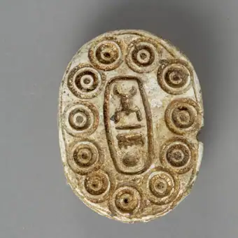 Amuletă sigiliu având partea superioară sculptată în forma unui scarabeu. Baza este decorată prin gravare cu următoarele elemente iconografice: în centru, într-un cadru rectangular patru hieroglife, i